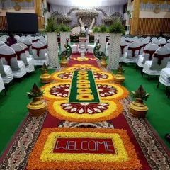 A welcoming floral rangoli at the entrance of a wedding hall. The design uses bright orange, yellow, and red flowers to create a vibrant path for guests, complete with a "Welcome" message.