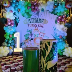 A close-up of a jungle-themed first birthday cake-cutting area. The setup includes a personalized 'Atharv Turns One' backdrop, a balloon arch with chrome and animal-print balloons, and themed pedestals.