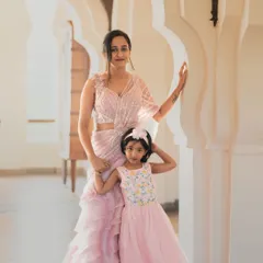 A mother and daughter in matching pastel pink outfits. The mother wears a modern ruffled saree, while her daughter is in an adorable matching gown, showing how I can create coordinated looks for all ages.