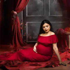 A serene, seated portrait from a fine art maternity session. The setup includes a floor covered in red fabric and petals, with dramatic curtains and florals in the background, creating a rich and romantic atmosphere.