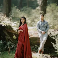A pre-wedding shoot in a sun-dappled forest. The bride's flowing red gown stands out beautifully against the green, creating a vibrant and romantic portrait.