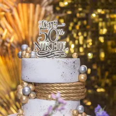 A close-up of the elegant two-tier cake for Nikhil's 50th birthday. The design features a textured white finish, a gold rope detail, and scattered gold and silver spheres.