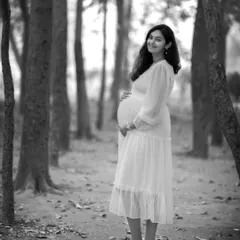 A stunning full-length solo portrait in black and white. The contrast between the white dress and the dark tree trunks creates a striking and artistic composition.