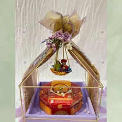 A beautiful Teej hamper featuring a Ghewar box placed inside a decorative metal cage adorned with a floral swing. The lavender color scheme adds a modern and elegant touch.