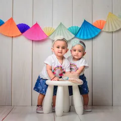 A colorful and fun studio session with twin girls. The bright paper fan garland and their matching rainbow shirts create a cheerful and vibrant atmosphere.