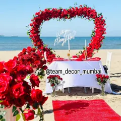 I design unforgettable marriage proposal setups, like this one on a Mumbai beach. It features a heart-shaped arch of red roses, a private dining table, and a 'Will you Marry Me?' sign, creating a picture perfect moment against the ocean backdrop.