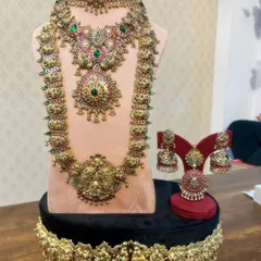This is a complete bridal set in antique gold finish with beautiful Kemp stones. It includes a choker, a long haram, matching jhumkas, a ring, and a vaddanam, everything a bride needs for her muhurtham.