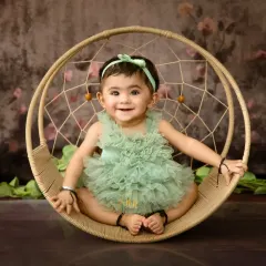 A happy baby girl in a green ruffled romper, sitting confidently in a round dreamcatcher swing.