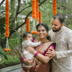 A lovely family portrait taken outdoors during a naming ceremony. The parents embrace their child under hanging marigold flower arrangements, creating a warm and traditional photograph.