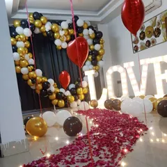 An indoor proposal setup designed for a grand surprise at home. This features a heart-shaped balloon arch in a classic black, gold, and white theme, large marquee "LOVE" letters, and a romantic walkway of scattered rose petals.