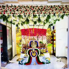 An elaborate floral mantap designed for a Varamahalakshmi puja at home. This decoration features a dense ceiling of white and red flowers flowing down into a multi-colored floral backdrop for the deity, creating a truly divine atmosphere.