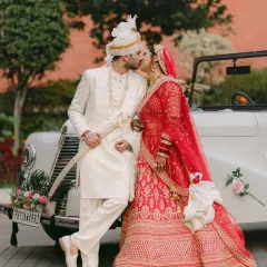 A moment of pure joy as the couple shares a kiss against a classic vintage car. The vibrant red of the bride's lehenga and the groom's elegant white sherwani create a cinematic portrait of their Punjabi wedding celebration.