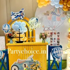 A hot air balloon and vintage vehicle theme for a first birthday. The setup features a bright yellow counter, a light-up number '1', and various props like airplanes and scooters.