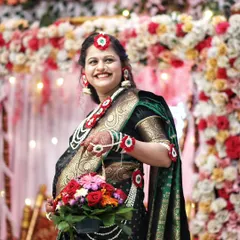 A radiant mother-to-be smiles brightly during her baby shower, holding a basket of fresh flowers. The vibrant floral decor in the background complements her traditional look.
