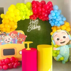 A vibrant and colorful Cocomelon-themed birthday party. The decor features a bright balloon arch over a faux grass wall, with large cutouts of JJ and other characters from the show.