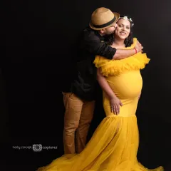 A joyful couple's maternity portrait against a dark studio background. The partner kisses the mom-to-be's cheek as she smiles in a vibrant yellow ruffled gown, showcasing a fun and loving interaction.