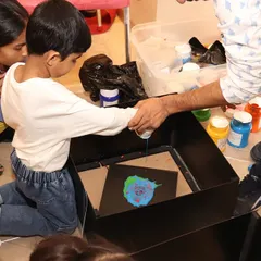 An instructor helps a young boy with a spin art project. By adding drops of paint to a spinning canvas, kids learn about centrifugal force while creating a unique masterpiece.