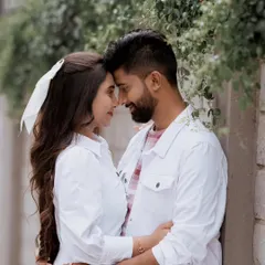 An intimate close-up of a couple in coordinated white outfits, sharing a quiet moment against a leafy urban wall.