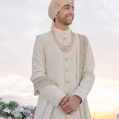 The groom's quiet moment of anticipation. Dressed in an elegant ivory sherwani, he smiles as he awaits his bride, a timeless portrait captured against the soft light of the sky.