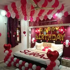 A classic romantic birthday bedroom setup. This decoration features red and white balloons on the ceiling and floor, "Happy Birthday" foil letters, and rose petals on the bed.