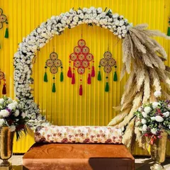 This is a popular setup for a Mehndi or Haldi ceremony. It combines a modern white floral arch with a traditional yellow backdrop and decorative hangings, creating a vibrant and stylish space for your pre-wedding functions.
