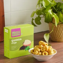 Quinoa rings are presented in a bowl next to their bright green packaging. The setting, complete with a houseplant, gives the snack a healthy, natural, and at-home feel.