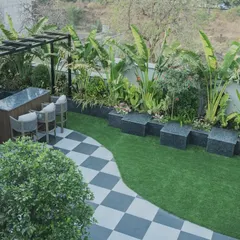 An overhead view of a terrace designed for both relaxation and entertainment. The layout includes a custom bar, a green lawn area, and checkerboard tiling, all surrounded by lush, tropical plants like the Traveller's Palm.