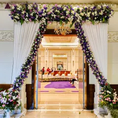 An elegant floral archway marks the entrance to the main hall. The combination of purple and white flowers with classic white drapes and a chandelier creates a sophisticated and welcoming feel.