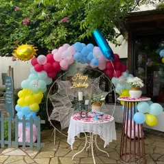 A colorful and cheerful birthday setup in a cafe's outdoor area. The decor includes a multi-colored balloon arch and a 'Happy Birthday' sign.