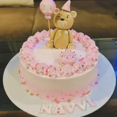 A sweet and simple first birthday cake in pink. It's topped with an adorable fondant teddy bear holding a "1" balloon, with the name "Navi" written in pink letters.