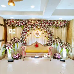 This engagement stage features a beautiful floral arch made of red, white, and purple blooms, with a custom initial monogram hanging in the center. The warm drop lighting adds a soft, romantic glow to the setting.
