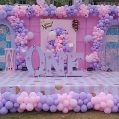 For this first birthday, we went all out with a grand stage setup. The design includes a beautiful pink and lavender balloon arrangement framing custom-built backdrops, with large 'ONE' letters and the birthday girl's name as the centerpiece.