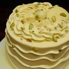 A close-up of another Rasmalai cake, this one with beautiful piped swirls on top. It's garnished with pistachios and real rasmalai pieces, promising a rich and flavorful experience.