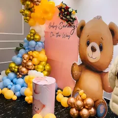 A cute teddy bear themed baby shower with a "Baby On The Way" message. This setup includes a large teddy bear cutout, a pink arched backdrop, and a colorful balloon arrangement in blue, yellow, and gold.