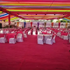 A large pandal setup with a colorful striped fabric ceiling and neatly arranged round tables with pink covers for a massive wedding lunch.