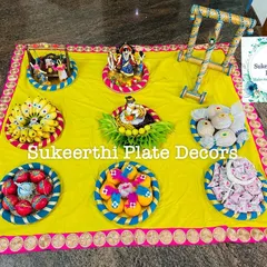 A colourful assortment of seer varisai plates on a bright yellow cloth, including a miniature walker, cradle, and other thematic items.