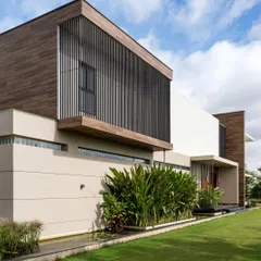 The home's exterior features strong geometric forms, with slatted screens providing both privacy and visual texture. The structure is designed to sit harmoniously within the surrounding mango plantations and cultivated land.