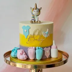 A "Oh Baby" unicorn-themed baby shower cake. The design combines a rainbow unicorn with classic baby elements like booties and onesies in a gender-neutral colour palette.