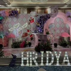 The main stage for the "Hridya's ONEderland" party. The setup is rich with details like a harlequin pattern backdrop, oversized foliage, marquee letters, and whimsical props.