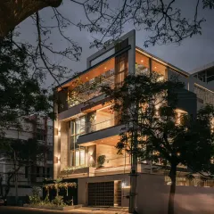 A corner view of the Hennur residence at night, framed by trees. This angle shows how the different volumes of the house come together, with the lighting scheme emphasizing the modern architectural form.