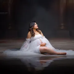 A dreamy and ethereal portrait of a mother-to-be in a white tulle gown. The reflective floor and moody lighting create a magical, water-like effect in the studio.