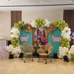 A complete Krishna-themed baby shower stage I created. This playful decor features a custom backdrop with Radha and Krishna, framed by an olive green and white balloon arch, and includes themed elements like cow cutouts, wicker chairs, and 'BABY' blocks for a perfect photo opportunity.