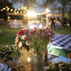 A close-up of a rustic table setting for a sundowner. A bouquet of wildflowers, a tall candle, and woven placemats create a warm and inviting atmosphere as evening sets in.