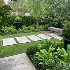 A tranquil garden path with stepping stones leading to a stone bench. The space is enclosed by a dense hedge and features large planters, creating a private and shaded seating area.