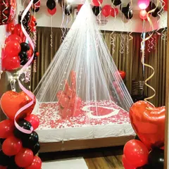 This romantic room decoration uses a classic red and black color scheme. The setup includes a fairy light canopy, a bed covered in rose petals, and heart-shaped foil balloons for a truly romantic surprise.