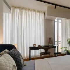 This bedroom corner is designed as a functional workspace, with a simple desk and chair positioned to receive ample natural light. Sheer white curtains diffuse the light, creating a bright yet private area.