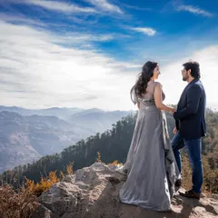 The couple stands on a rocky outcrop, holding hands and looking out at the vast blue sky and mountain vista. This is a classic adventurous pre-wedding shot.