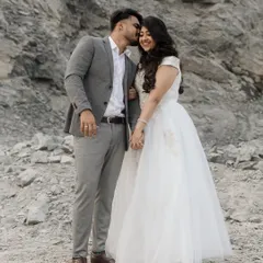 A sweet and affectionate moment against a rugged, natural backdrop. He kisses her on the cheek as she smiles, holding his hand. This full-length shot captures their joy and the beautiful contrast of her white dress against the rocks.