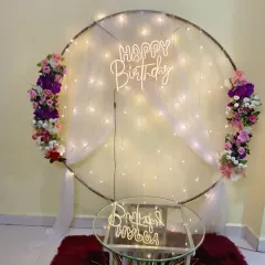 A circular frame with fairy lights and white drapes serves as a simple and elegant backdrop. The floral accents and "Happy Birthday" neon sign complete the look.