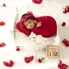 A beautiful newborn girl in a red wrap and floral headband, sleeping on a white bed with a calendar block marking the date.
